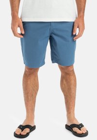 Blå shorts i lätt tyg med rak framdel, sidofickor och en avslappnad passform, tillsammans med svarta flip-flops.