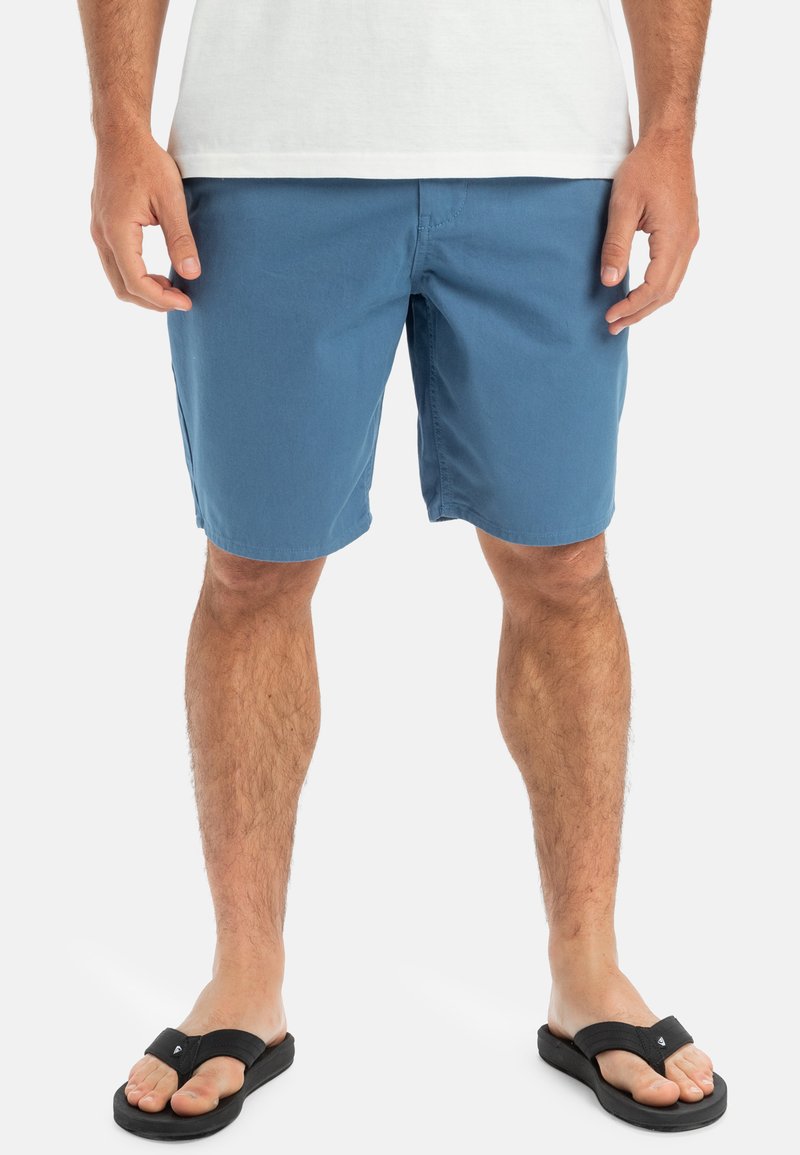 Blå shorts i lätt tyg med rak framdel, sidofickor och en avslappnad passform, tillsammans med svarta flip-flops.