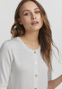 Hvid strikket cardigan med rund halsåbning, trekvartærmer og dekorativ knaplukning. Glat tekstur, let og blødt materiale.
