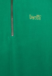 Suéter de forro polar verde con cremallera de un cuarto de longitud y una textura suave. Presenta un logo bordado amarillo en el pecho.