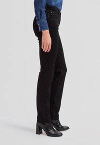 Persoană purtând pantaloni negri skinny, cizme negre cu toc la gleznă și o cămașă albastră cu mânecă lungă și nasturi, stând pe o parte.