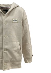 Felpa leggera beige con chiusura frontale a bottoni, ampie tasche, polsini a coste e una toppa con logo sul petto. Tessuto morbido.