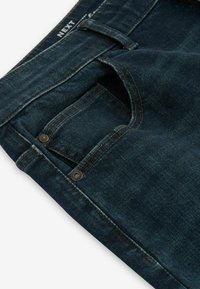 Tmavě modré džíny z denimového materiálu s viditelným šitím, přední kapsou a kovovými akcenty. Látka má hladkou, mírně opotřebovanou strukturu.