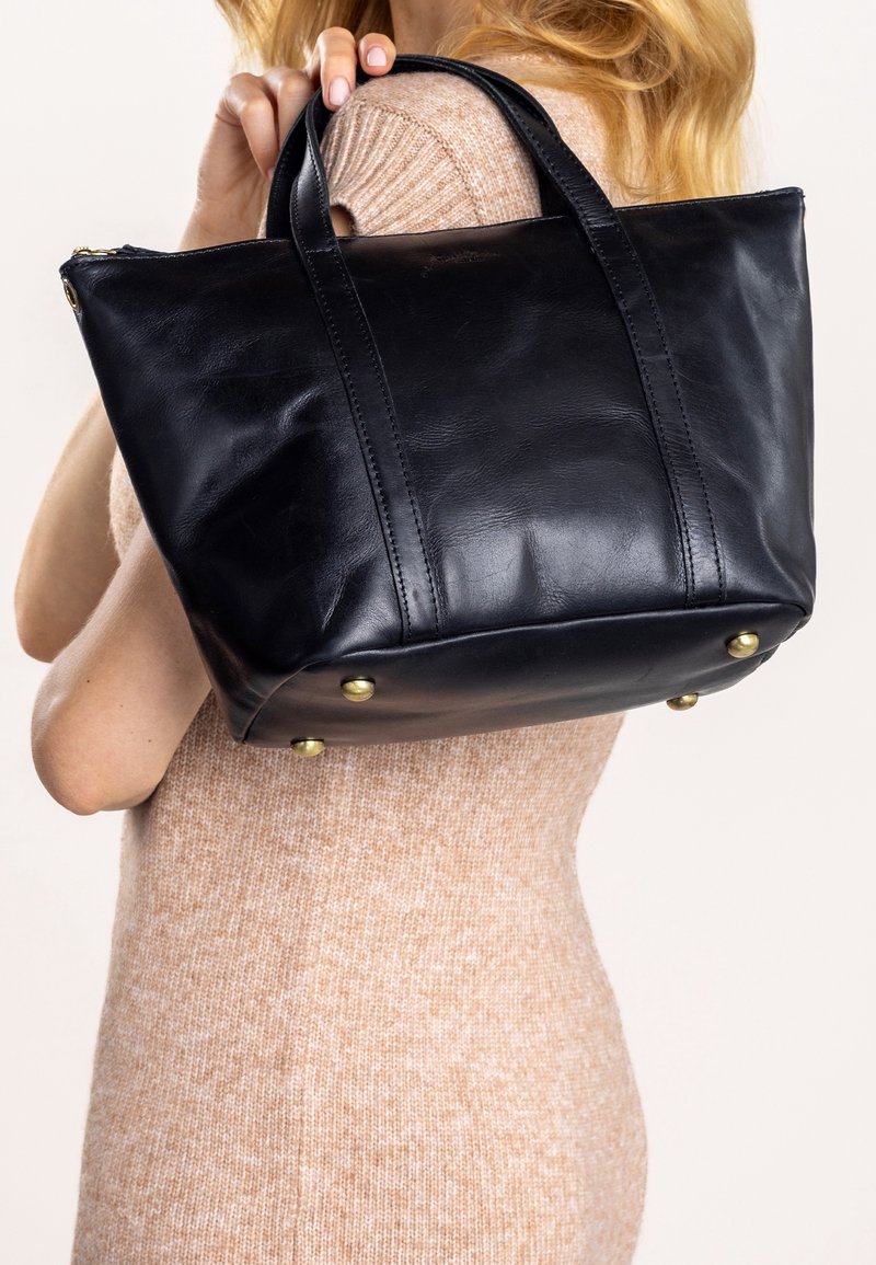 Femme tenant un sac fourre-tout en cuir noir avec des clous dorés en bas, portant une robe en maille beige, vue de dos et de profil.