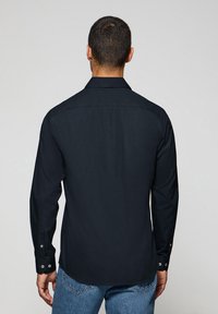 Camisa negra de manga larga con puños abotonados, dobladillo recto y detalles en el yugo trasero. Tejido liso sin patrones visibles. Ajuste casual.