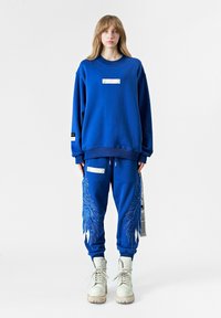 Sudadera azul con una etiqueta en el pecho, combinada con pantalones azules que tienen bordados de alas. Botas blancas completan el conjunto.