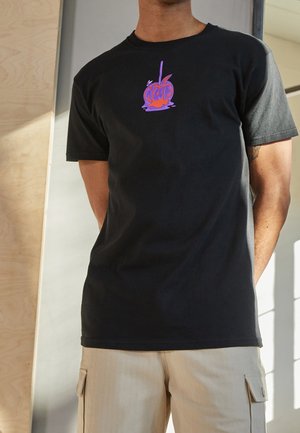 Personne portant un t-shirt noir avec un motif de pomme d'amour violette et orange et un pantalon cargo beige, debout à l'intérieur, les bras derrière le dos.