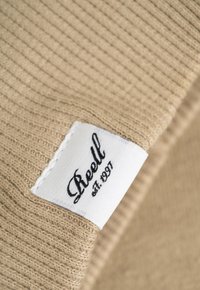 Beige gestrickter Stoff mit gerippter Textur; enthält ein weißes Etikett, das den Markennamen "Reell" und das Gründungsjahr "est. 1997" zeigt.