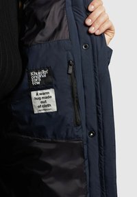 Marine pufferjack binnenkant met ritsvak en twee labels. Materiaal is zacht en gewatteerd, met zwart-witte tekst op de labels.