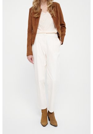 Femme debout portant une veste en daim marron, un chemisier crème, un pantalon taille haute blanc et des bottines en daim marron sur fond blanc.