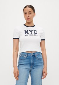 Hvit cropped t-skjorte med marineblå ribbestrikket krage, med fet tekst "NYC" og "NEW YORK CITY" i marineblått. Kombinert med lyseblå jeans.