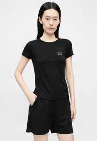 Tricou negru cu mânecă scurtă din material moale, având un mic logo EA7 într-o culoare contrastantă pe piept. Modelul poartă pantaloni scurți negri.