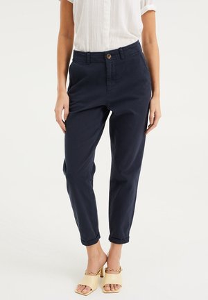 Kobieta ubrana w granatowe, wysokowciskie spodnie zwężane z mankietami u dołu, w połączeniu z beżowymi sandałami na obcasie z detalem łańcuszka.