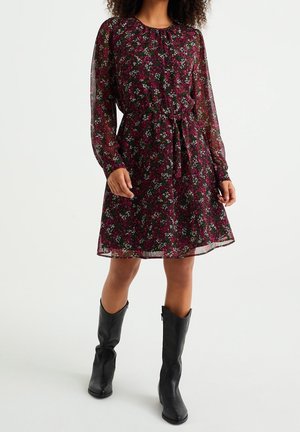 Femme portant une robe à manches longues, longueur genoux, à motif floral en rouge foncé et noir avec une ceinture à la taille, associée à des bottes noires mi-mollet.