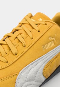 Gelbe Wildleder-Sneaker mit einem weißen Lederakzent, flachen Schnürsenkeln und einem goldfarbenen Logo. Verfügt über strukturierte Seitenpaneele und eine schwarze Gummisohle.