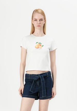 Cropped top bianco a maniche corte con stampa di frutti arancioni, abbinato a pantaloni shorts in denim scuro con vita alta e fit rilassato.