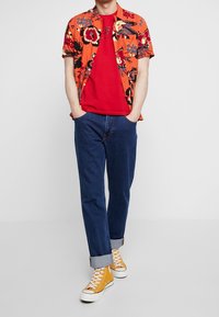 Kortærmet orange blomstret skjorte over en rød T-shirt, parret med opsmøgede blå jeans og gule høj-top sneakers.