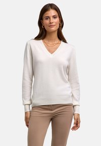Donna che indossa un maglione bianco a V, pantaloni beige, orecchini a cerchio dorati e una sottile collana d'oro, in piedi davanti a uno sfondo semplice.