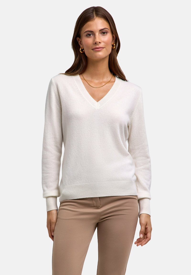 Donna che indossa un maglione bianco a V, pantaloni beige, orecchini a cerchio dorati e una sottile collana d'oro, in piedi davanti a uno sfondo semplice.