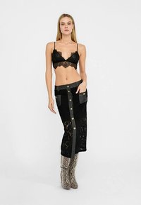 Zwarte kanten bralette gecombineerd met een lange zwarte kanten rok met metalen knoppen en twee zijzakken, gedragen met zilveren laarzen met slangprint.