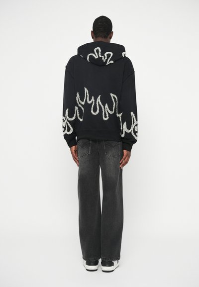 Άτομο με μαύρη κουκούλα με λευκό μοτίβο φλόγας και χαλαρούς σκούρους γκρι τζιν, που στέκεται μπροστά σε λευκό φόντο.