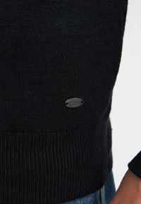 Schwarzer Strickpullover mit Rippstrick am Saum, ausgestattet mit einem kleinen silbernen Logo-Patch mit der Bezeichnung "Petrol", das an der unteren Seite angenäht ist.