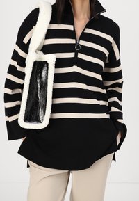 Suéter a rayas negras y crema con cuello de cremallera hasta el pecho. Combinado con un forro polar blanco texturizado y un bolso negro brillante. Pantalones neutros.