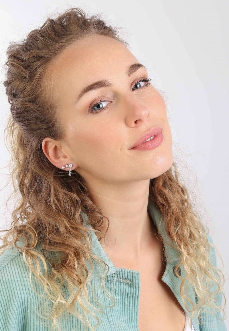 Zilveren studs met verschillende vormen, versierd met sprankelende stenen, gedragen door een persoon met krullend haar en een lichtblauwe geribbelde jas.