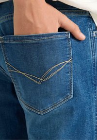 Tascone di jeans in denim blu con una mano parzialmente visibile. La tasca presenta cuciture chiare a contrasto disposte in un motivo a V decorativo.