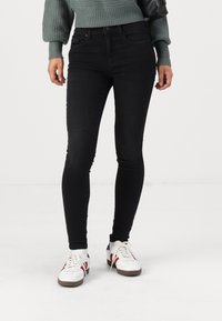 Black skinny jeans made of denim, featuring a high waist and a smooth texture. Paired with white sneakers and a green knitted sweater.