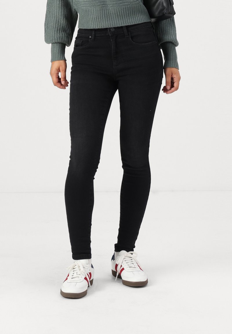 Black skinny jeans made of denim, featuring a high waist and a smooth texture. Paired with white sneakers and a green knitted sweater.