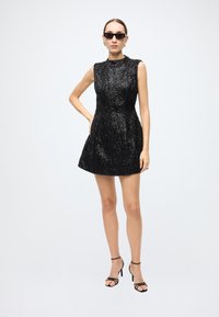 Black sequin dress with a high neckline and sleeveless design, flaring at the skirt. Accompanied by black strappy heels and sunglasses.