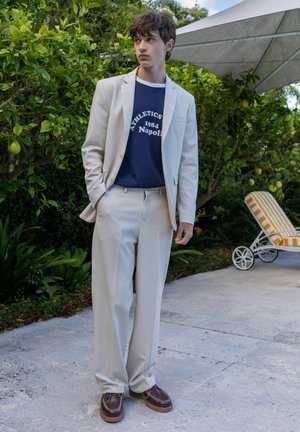 Jeune homme portant un costume beige et un t-shirt bleu marine « Athletics 1984 Napoli » debout à l'extérieur près de citronniers et d'une chaise longue rayée jaune.