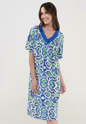 Mujer sonriente con cabello largo y castaño, vestida con un vestido hasta la rodilla con patrones abstractos en azul, verde y blanco, y un cuello en V sólido azul.