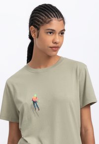 Licht olijfgroene katoenen t-shirt met een kleurrijk gedrukt ontwerp van een figuur in beweging op de voorkant. Korte mouwen, ronde halslijn.