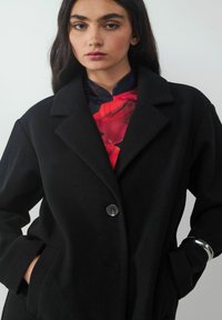 Schwarzer Wollmantel mit Reverskragen, einem einzelnen Knopfverschluss und Seitentaschen. getragen über einer rotblumigen Bluse. Einfaches und elegantes Design.