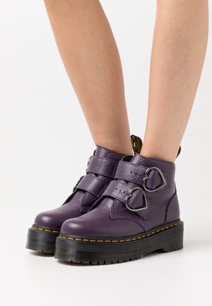 Bottines en cuir violet avec semelles noires épaisses et deux boucles métalliques en forme de cœur, portées sur des jambes nues.