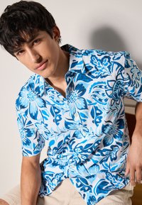 Chemise à manches courtes en tissu blanc ornée d'un motif floral bleu. Design boutonné avec une texture douce, mettant en valeur différentes nuances de bleu.