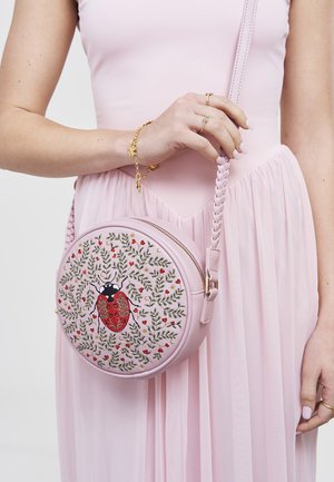 Vrouw in een roze jurk die een ronde roze handtas vasthoudt met een geborduurd ontwerp van een lieveheersbeestje en een bladerrank, met een gevlochten band over de schouder.