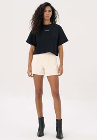 Zwarte cropped t-shirt met witte logo, gecombineerd met crèmekleurige shorts met een rafelrand, en zwarte enkellaarsjes. Eenvoudig, casual ontwerp.