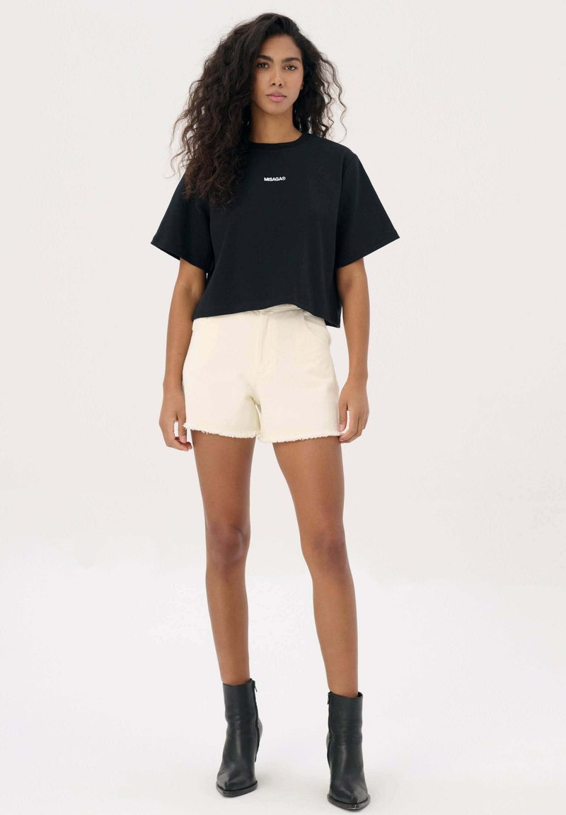 Zwarte cropped t-shirt met witte logo, gecombineerd met crèmekleurige shorts met een rafelrand, en zwarte enkellaarsjes. Eenvoudig, casual ontwerp.