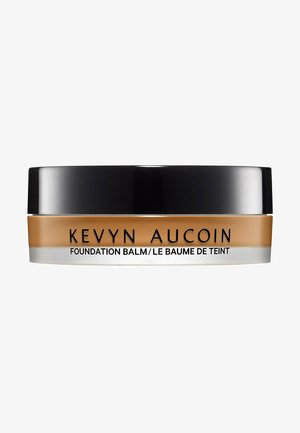 Kevyn Aucoin Foundation Balm in einem runden, schwarzen und klaren Behälter. Cremige Textur; goldbraune Farbe; Text auf dem Etikett in Schwarz.