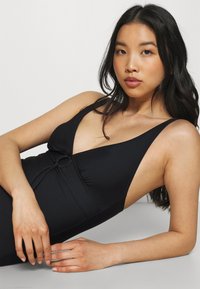 Zwart badpak met een diepe halslijn, verstelbare schouderbanden en een vooraan geknoopt detail. Het gladde, rekbare materiaal zorgt voor comfort en een goede pasvorm.
