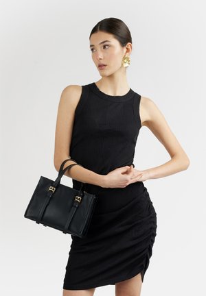 Donna che indossa un vestito nero senza maniche e tiene una borsa nera, con orecchini dorati a forma di fiore, su sfondo chiaro e uniforme.