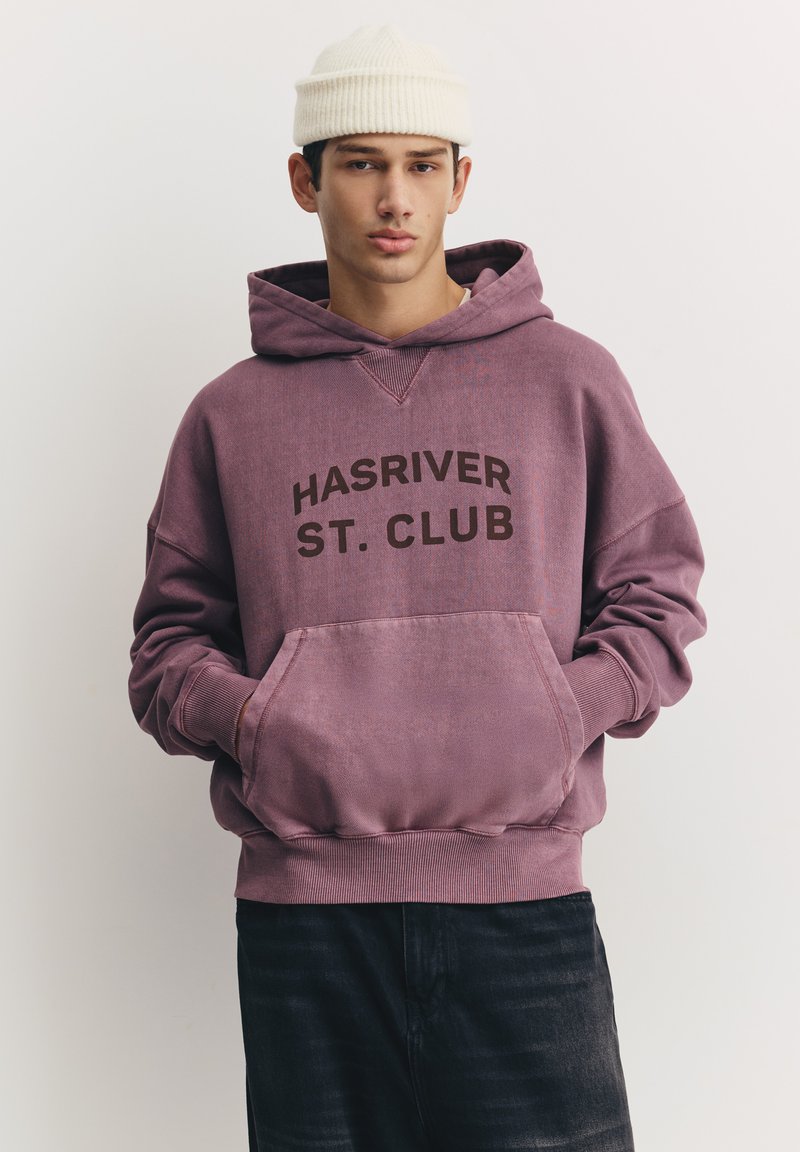 Sweat à capuche en coton violet avec une poche frontale, arborant l'inscription "HASRIVER ST. CLUB" en lettres brunes épaisses. Poignets et ourlet côtelés. Porté avec un pantalon foncé.