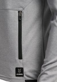 Grijze jas van gestructureerde stof, met een zwarte ritssluiting pocket en een logopatch met een chevronontwerp op de zoom.