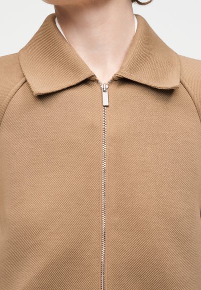 Άτομο που φοράει μπεζ τζάκετ από ριμπ ύφασμα με ασημένιο φερμουάρ κλεισμένο μέχρι τον γιακά.