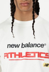 Balts kokvilnas T-krekls ar drosmīgu melnu un sarkanu tekstu "new balance ATHLETICS". Ietver dzeltenu New Balance logo etiķeti. Ar apaļu kaklu.