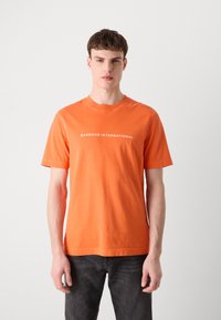 Oranžové bavlněné tričko s krátkými rukávy a kulatým výstřihem. Na přední straně má bílé nápisy "BARBOUR INTERNATIONAL".