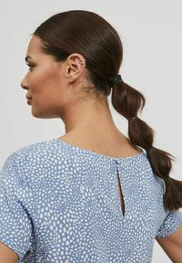 Blusa azul con patrón de puntos blancos texturizados, mangas cortas y un detalle de abertura en la parte posterior. Cabello recolectado en una coleta baja, sujetado con una banda negra.